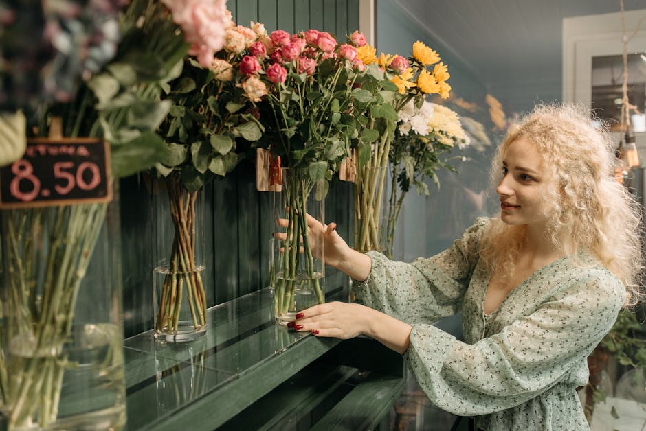 A woman with curly blonde hair, dressed in a light green floral blouse, stands inside a flower shop selecting a bouquet of fresh flowers. She is holding a tall, clear glass vase filled with pink roses