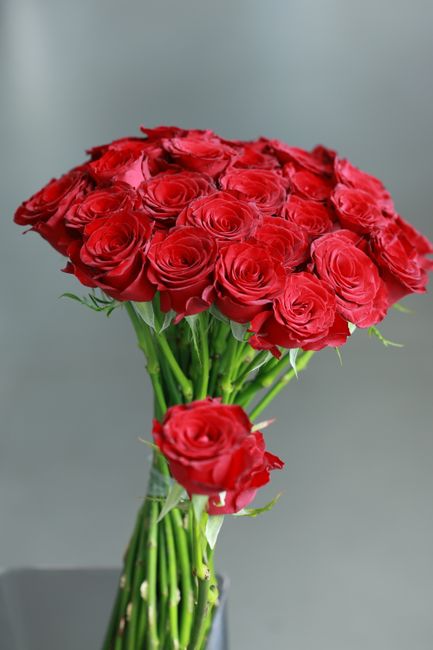 A bouquet of fresh red roses with tightly packed, velvety petals arranged in a rounded cluster, held together by green stems and leaves. The bouquet is displayed in a clear glass vase, showing the lon